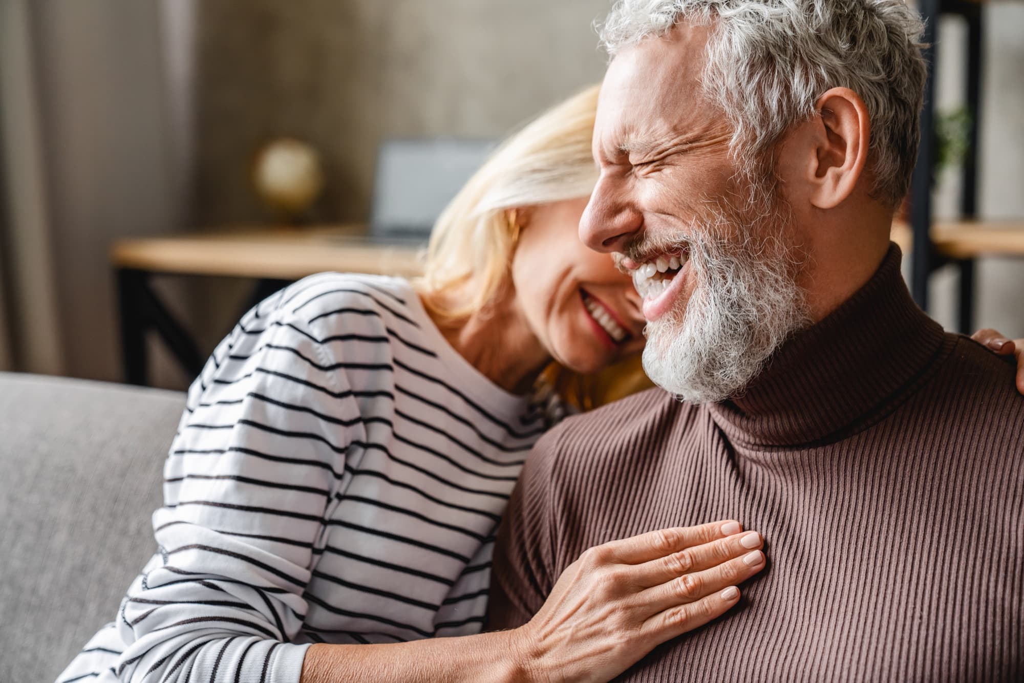 An older couple laughing together on a sofa, her head resting on his shoulder